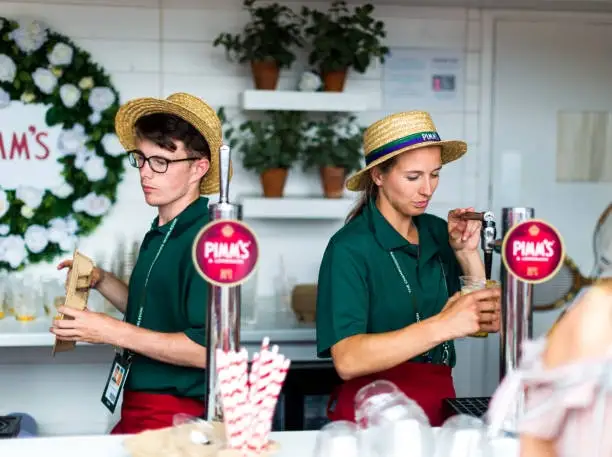 London, UK - 10 July, 2018: color image depicting two young bartenders, a young man and a young woman, wearing straw boater hats and serving Pimm's on tap behind a pop-up drinks bar outdoors on the south bank in London, UK. Focus is on the Pimm's taps in the foreground. Room for copy space.