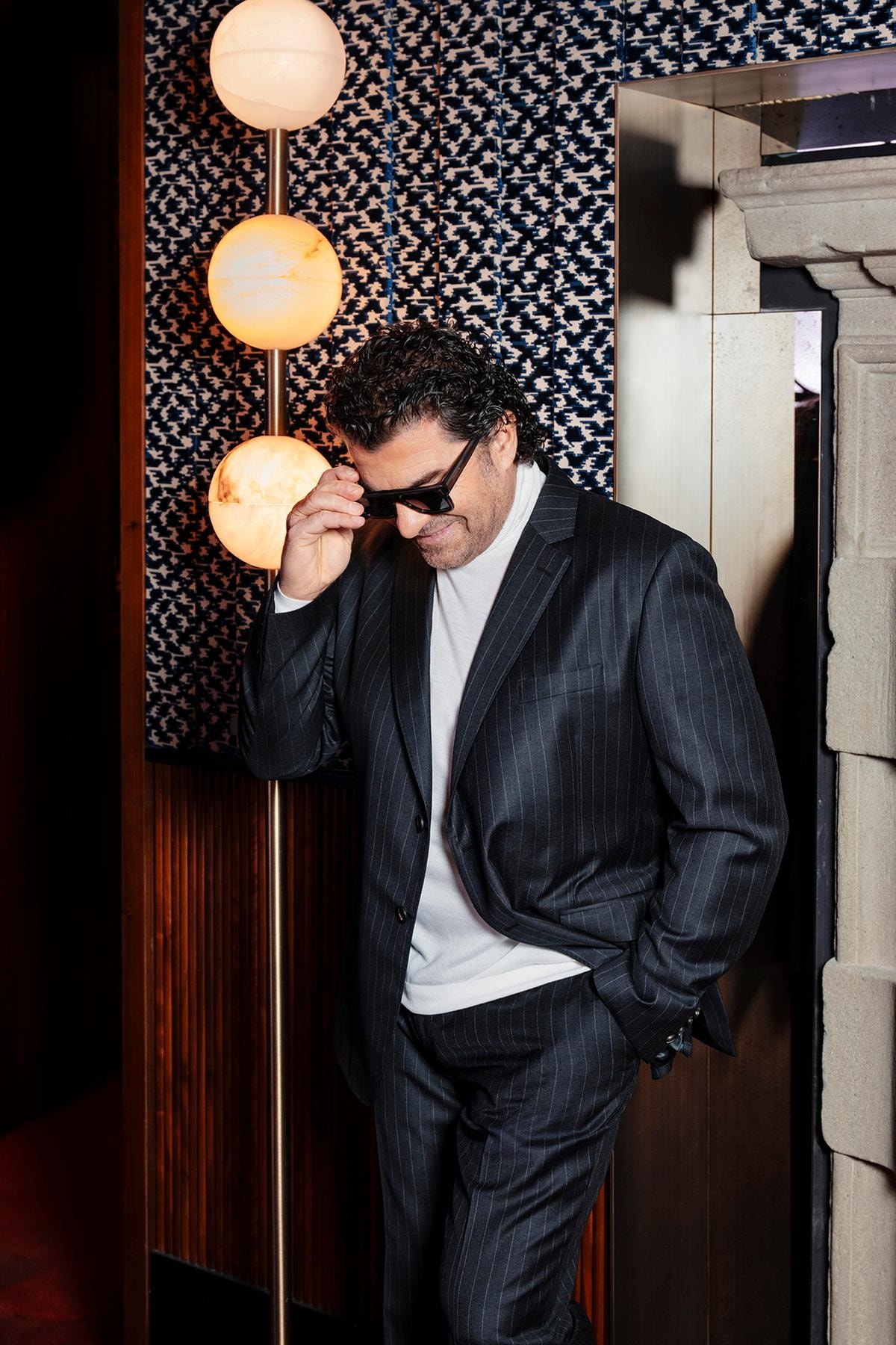 Alberto Tomba indossa un abito sartoriale in pura lana in tessuto gessato grigio, lupetto in lana e seta bianco, scarpe nere stringate con costruzione Tramezza in vitello semi lucido e occhiali da sole, tutto di Ferragamo.