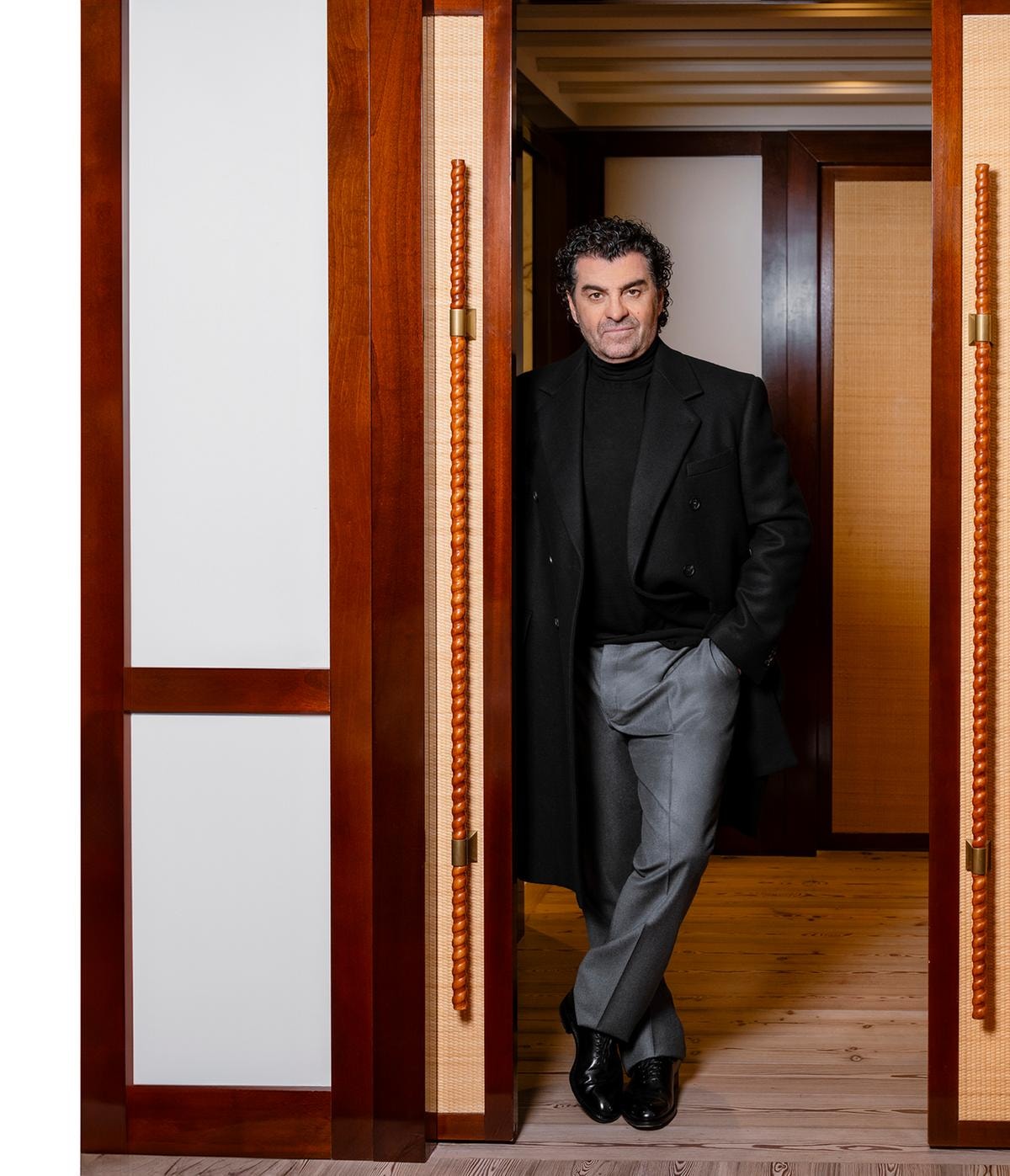 Alberto Tomba indossa un cappotto sartoriale nero in panno di lana con chiusura doppiopetto e revers, lupetto in lana e seta nero e scarpe nere stringate con costruzione Tramezza in vitello semi lucido, tutto di Ferragamo.
