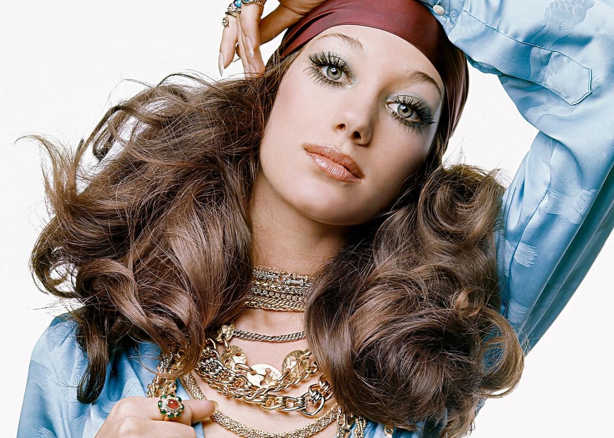 Model, Marisa Berenson, looks at the camera, her arm resting on the top of her head; she wears a long sleeve turquoise shirt by Capucci which is tied at front, with many gold chains and gold rings by Bulgari; a purple scarf is wrapped around her head; her dramatic blue eyeshadow is by Elizabeth Arden