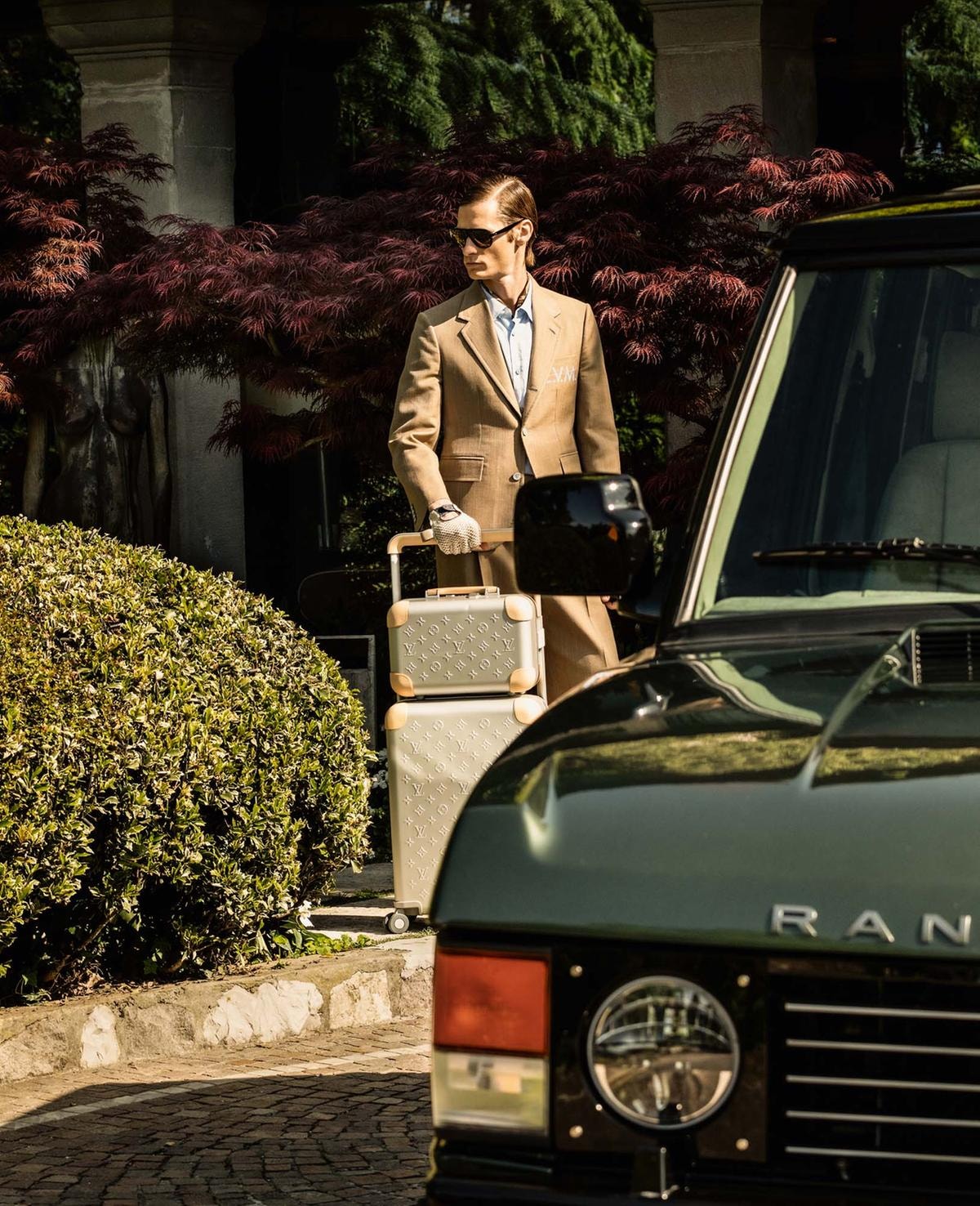Abito con giacca due bottoni  in lana e seta, camicia  in cotone, e foulard in seta, tutto di Louis Vuitton; occhiali da sole della collezione Paul Newman  di Oliver Peoples; guanti  da guida in pelle e cotone, Vinile per Restelli; trolley Horizon in alluminio e Vanity Case in alluminio con inserti in pelle, di Louis Vuitton.