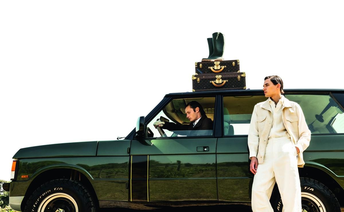 In primo piano, gilet con tasche applicate, camicia morbida, pantaloni e cache-col in lino, tutto di di Loro Piana. Seduto sulla Range Rover Classic by Vinile, sahariana in lino, camicia in cotone, pantalone in lino, tutto Kiton; guanti in pelle e cotone, Vinile per Restelli. Sopra, set di valigie in tela monogram con angoli e chiusura in ottone dorato, Louis Vuitton; stivali in gomma, Hunter.